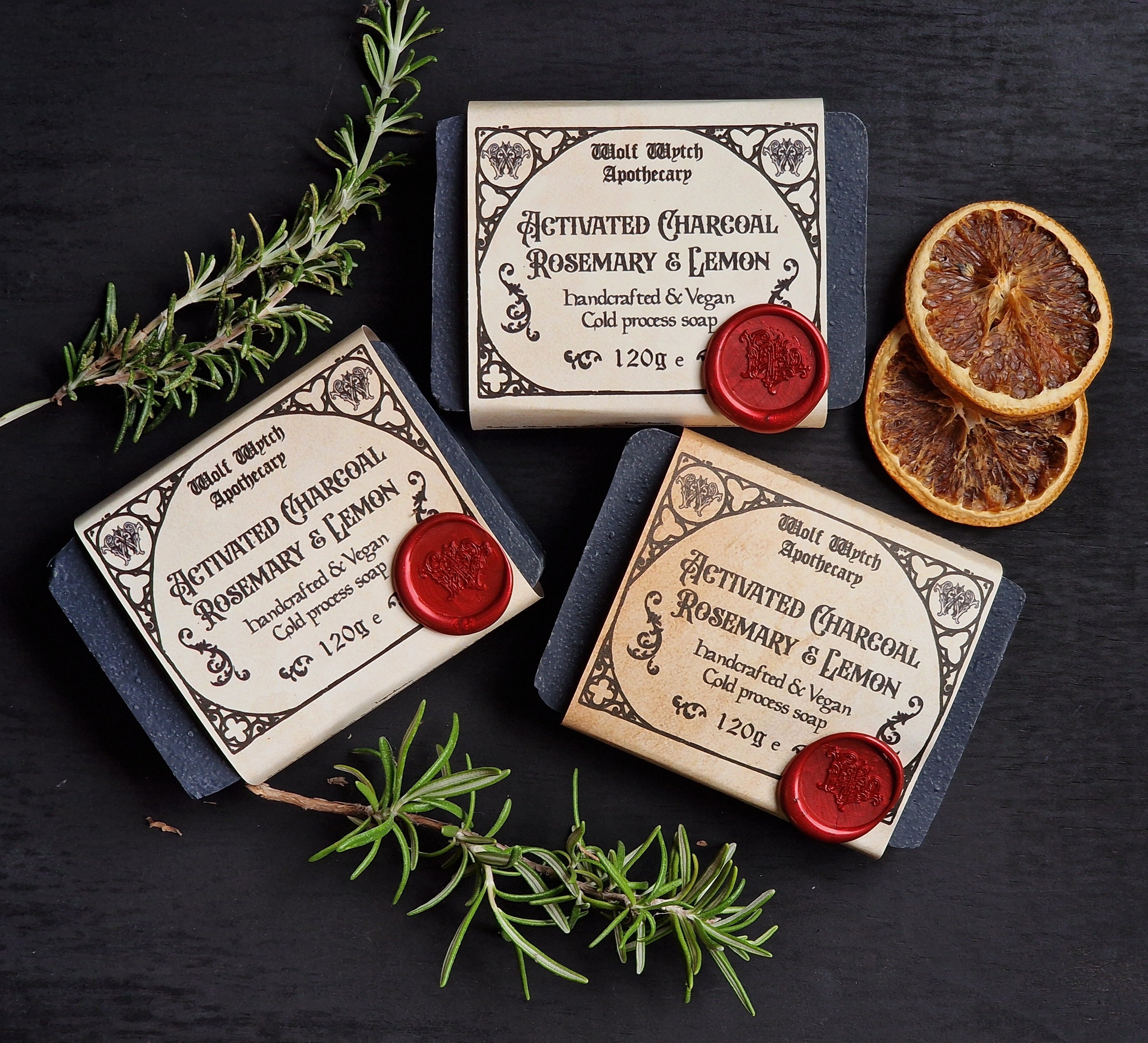 Three bars of soap with labels and red wax seals on a dark surface with rosemary and lemon slices.