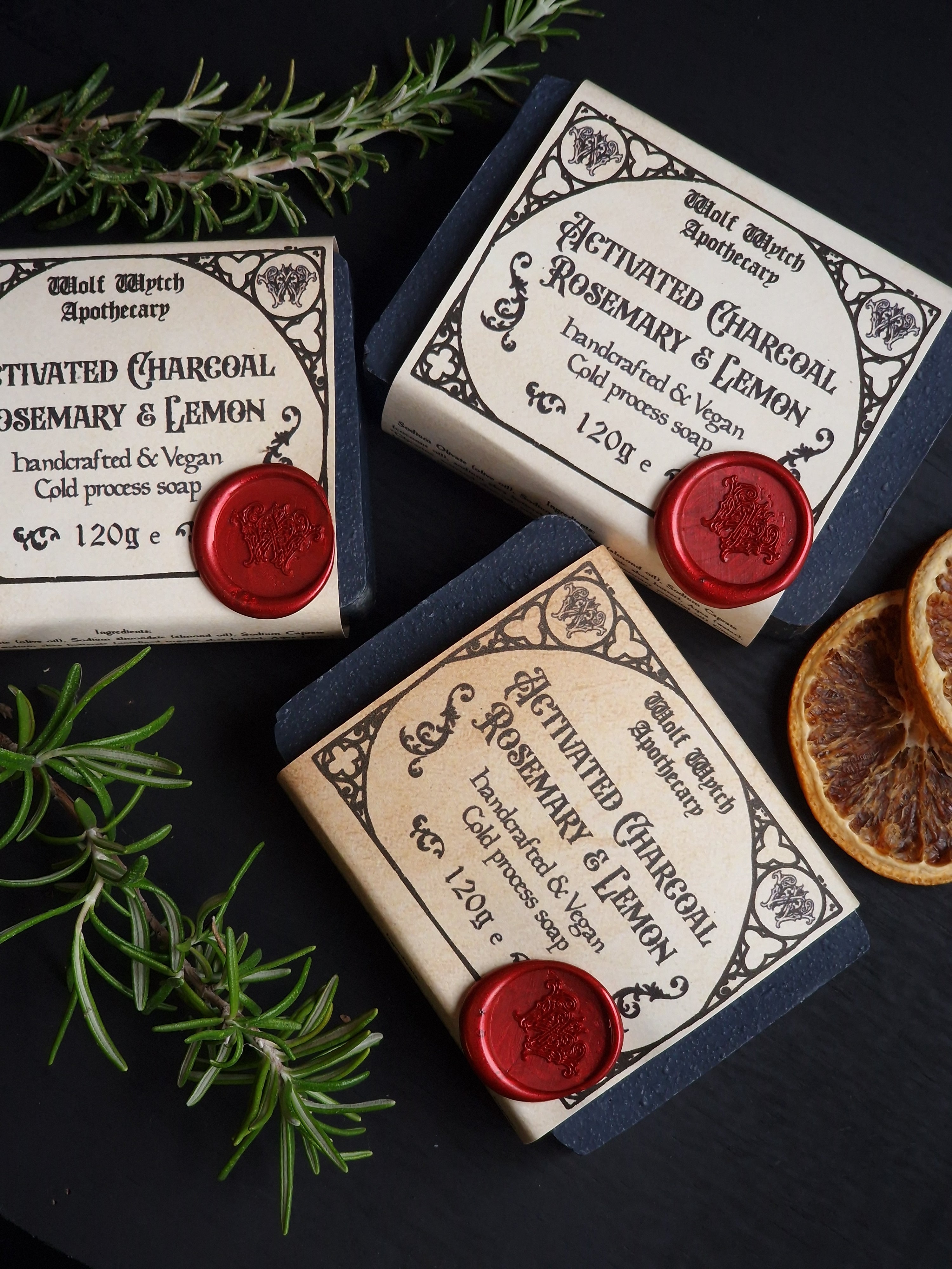 Three soap bars with decorative labels and red wax seals on a dark surface with rosemary sprigs.