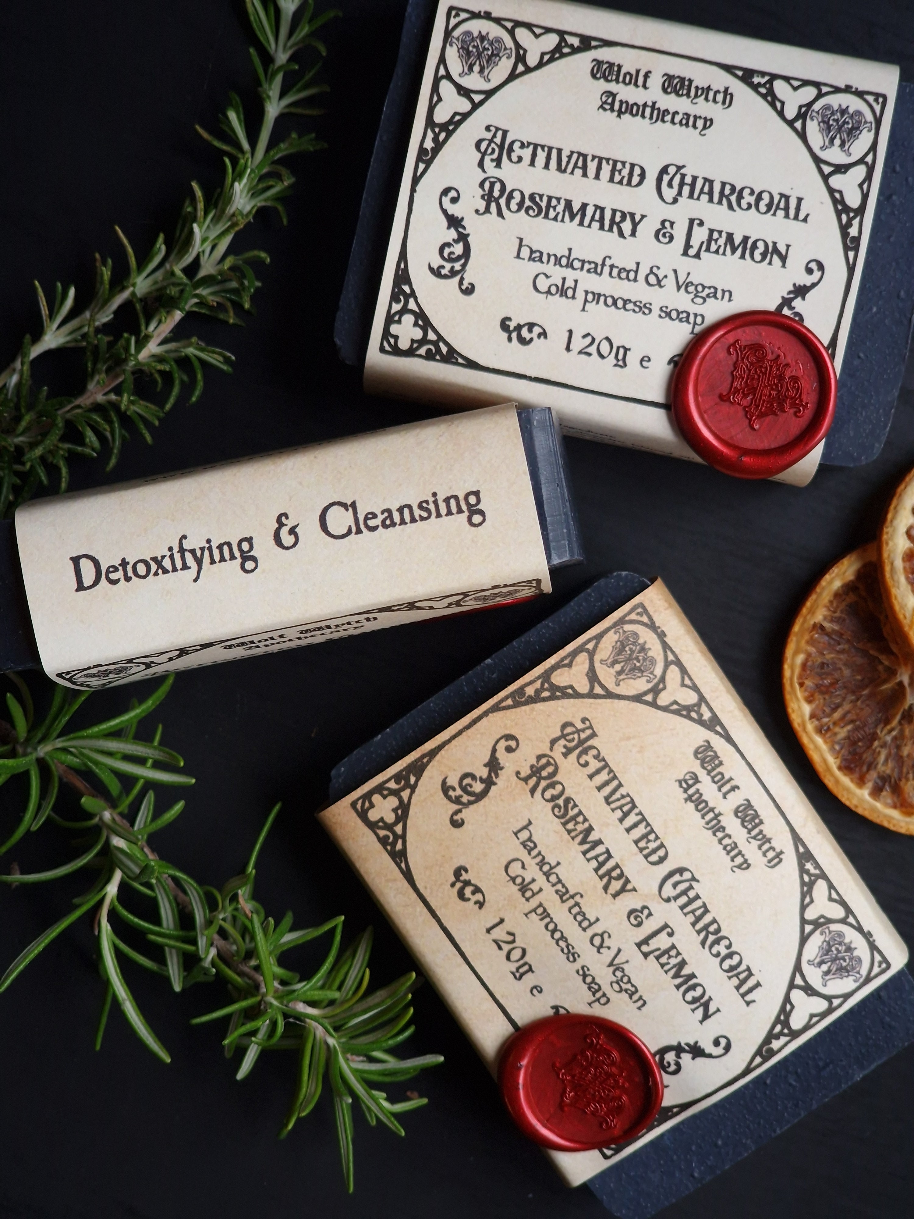 Two soap bars with labels and a red wax seal on a dark surface with herbs.