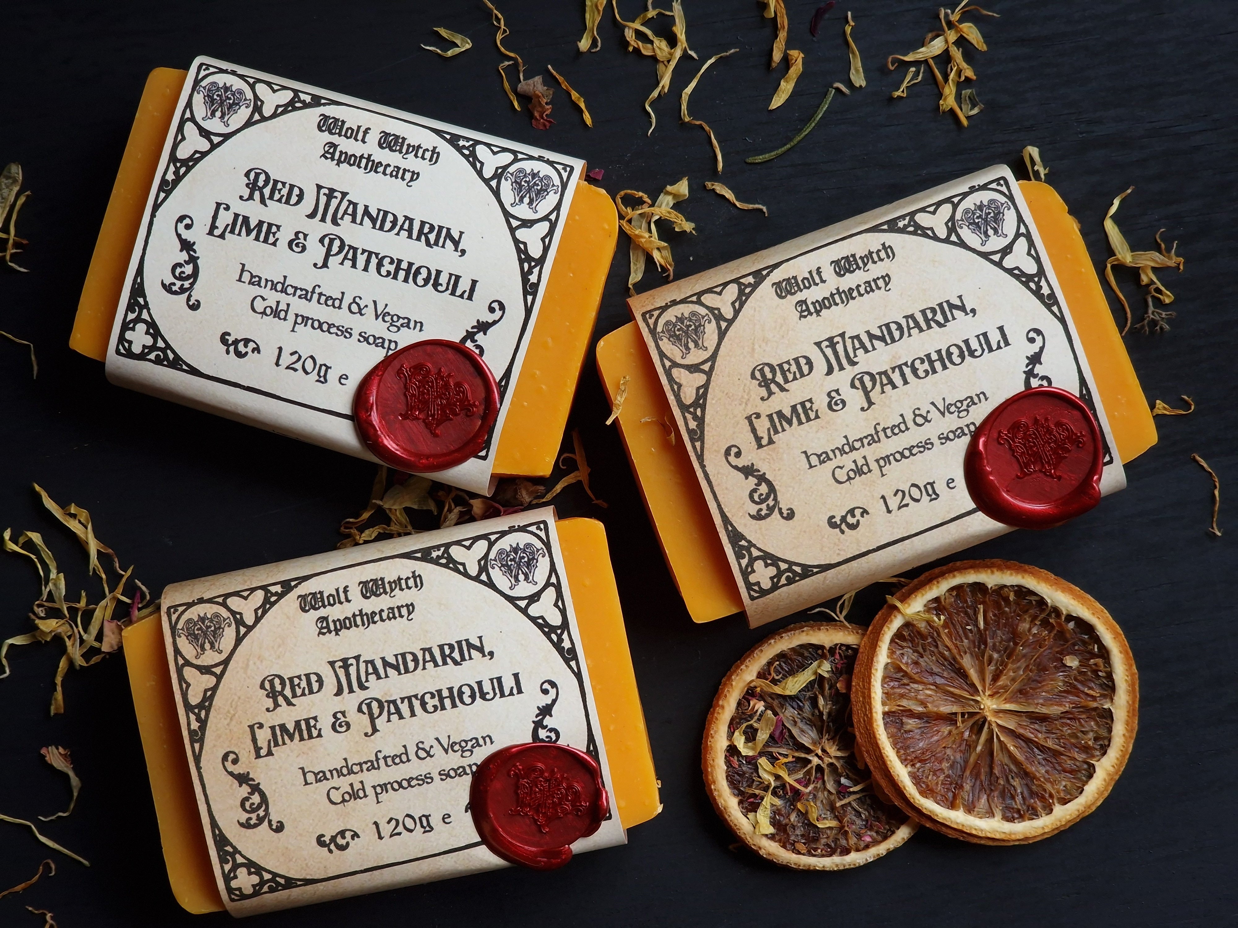 Three soap bars with decorative packaging on a dark surface with dried oranges and leaves.