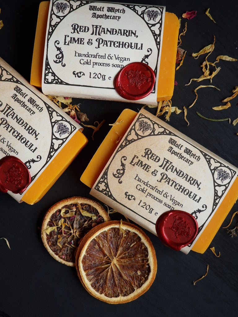 Three soap bars with decorative packaging and red wax seals on a dark surface with dried oranges.