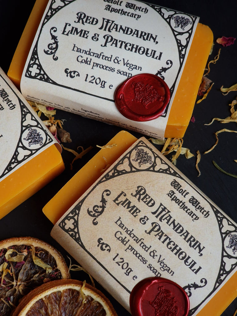 Three soap bars with decorative labels and red wax seals on a dark surface with dried oranges and leaves.