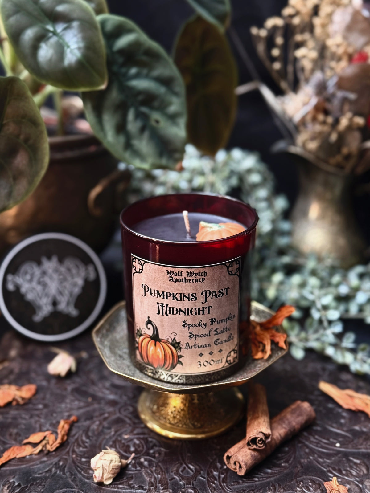 A dark amber glass candle in a decorative pedestal holder, labeled 'Pumpkins Past Midnight' by Wolf Witch Apothecary. The candle is styled with dried leaves, cinnamon sticks, and green foliage in the background.